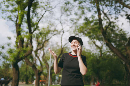 Young athletic smiling beautiful brunette girl in black uniform and cap talking on mobile phone during training, looking aside and standing in city park outdoors. Fitness, healthy lifestyle conceptの写真素材