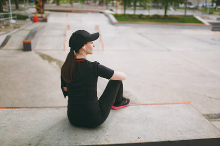 Back view Young athletic brunette girl in black uniform and cap with headphones listening music resting and sitting before or after running, training in city park outdoors. Fitness, healthy lifestyleの写真素材