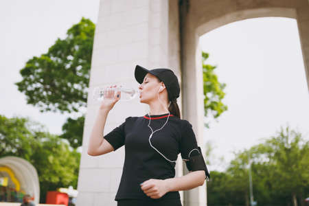 Young athletic beautiful brunette girl in black uniform, cap with headphones holding bottle drinking water during training listening music standing in city park outdoors. Fitness, healthy lifestyleの写真素材
