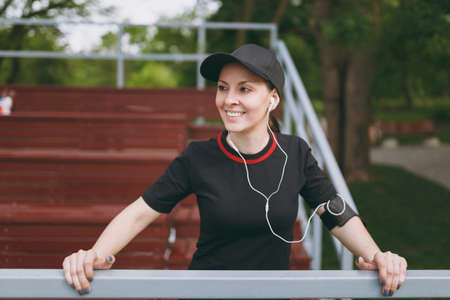 Young athletic smiling girl in black uniform and cap with headphones listening music, resting and...