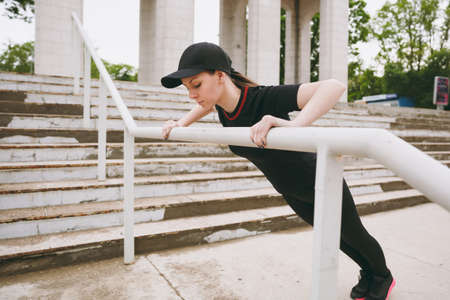 Young concentrated athletic beautiful brunette girl in black uniform and cap doing sport exercises, making push-up warm-up before running in city park outdoors. Fitness, healthy lifestyle conceptの写真素材