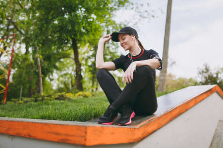 Young athletic relaxing beautiful girl in black uniform and cap with headphones listening music resting and sitting before or after running, training in city park outdoors. Fitness, healthy lifestyleの写真素材