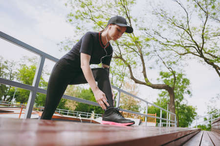 Young concentrated athletic brunette girl in black uniform, cap with headphones listening music doing sport stretching exercises warming-up on bench in city park outdoors. Fitness, healthy lifestyleの写真素材