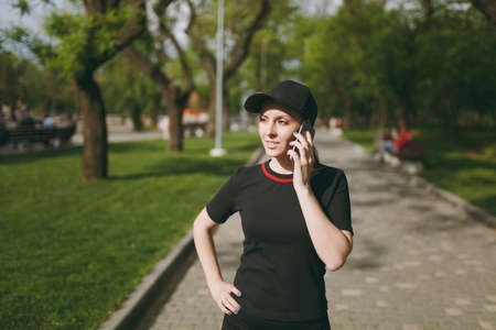 Young athletic beautiful brunette girl in black uniform and cap talking on mobile phone during training, looking aside and standing in city park outdoors. Fitness, healthy lifestyle conceptの写真素材