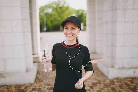Young athletic smiling beautiful brunette girl in black uniform and cap with headphones holding bottle with water during training, listening music in city park outdoors. Fitness, healthy lifestyleの写真素材