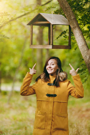 Beautiful happy caucasian young smiling brown-hair woman in yellow coat, jeans, boots in green forest. Fashion model with fall leaves standing under bird feeders in early autumn park outdoorsの写真素材