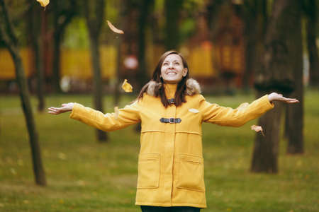 Beautiful happy caucasian young smiling brown-hair woman in yellow coat, jeans, boots in green forest. Fashion female model throwing up fall leaves standing and walking in early autumn park outdoorsの写真素材