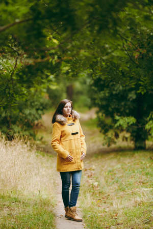 Beautiful happy caucasian young smiling brown-hair woman in yellow coat, jeans, boots in green forest. Fashion female model with fall golden leaves standing and walking in early autumn park outdoorsの写真素材