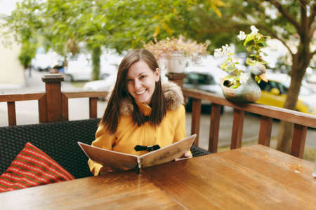 Beautiful happy caucasian young smiling brown-hair woman in yellow coat reading, ordering from menu in outdoor restaurant or cafe near road and deciding what to eat on dinner in autumn seasonの写真素材
