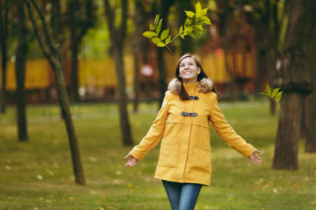Beautiful happy caucasian young smiling brown-hair woman in yellow coat, jeans, boots in green...