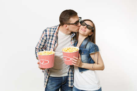 Young happy couple, woman and man in 3d glasses and casual clothes watching movie film on date holding buckets of popcorn, hugging and kissing isolated on white background. Emotions in cinema conceptの写真素材