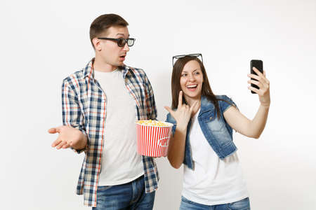 Young couple, woman and man in 3d glasses watching movie film on date holding bucket of popcorn doing selfie on mobile phone, showing rock-n-roll sign isolated on white background. Emotions in cinemaの写真素材