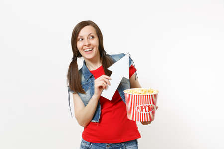 Portrait of young smiling woman in casual clothes watching movie film, holding bucket of popcorn, pointing white arrow up on copyspace isolated on white background. Emotions in cinema conceptの写真素材