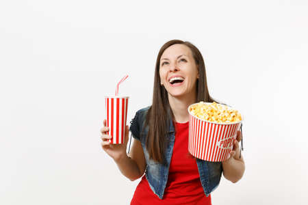 Portrait of young funny laughing attractive woman watching movie film, holding bucket of popcorn and plastic cup of soda or cola, looking up isolated on white background. Emotions in cinema conceptの写真素材