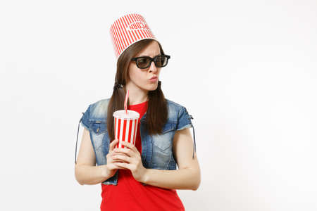 Portrait of young concerned pretty woman in 3d glasses with bucket for popcorn on head watching movie film and holding plastic cup of soda or cola isolated on white background. Emotions in cinemaの写真素材