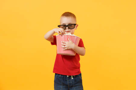 Little cute kid baby boy 3-4 years old in red t-shirt, 3d cinema glasses holding bucket for popcorn, eating fast food isolated on yellow background. Kids childhood lifestyle concept. Copy spaceの写真素材