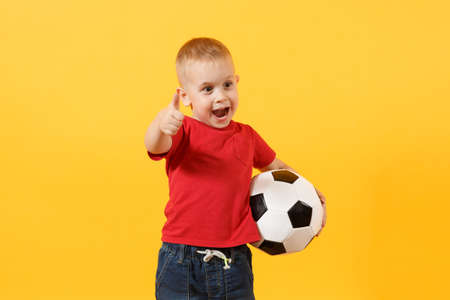Little cute kid baby boy 3-4 years old, football fan in red t-shirt holding in hand soccer ball isolated on yellow background. Kids sport family leisure lifestyle concept. Copy space advertisementの写真素材