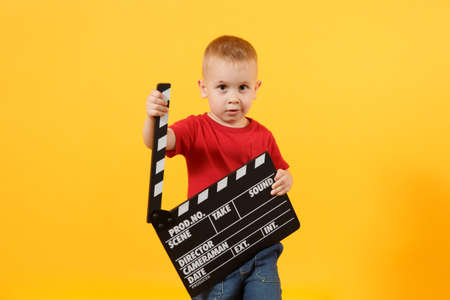 Little cute kid baby boy 3-4 years old in red t-shirt holding in hand classik black film making clapperboard looking camera isolated on yellow background. Kids childhood lifestyle concept. Copy spaceの写真素材