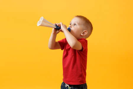 Little cute kid baby boy 3-4 years old, football fan in red t-shirt holding in hand pipe, blowing isolated on yellow background. Kids sport family leisure lifestyle concept. Copy space advertisementの写真素材
