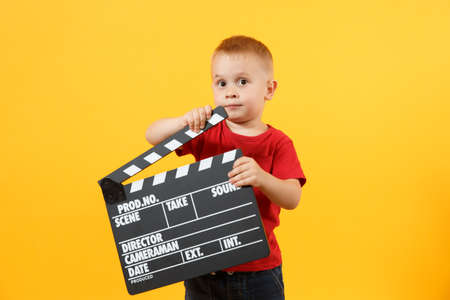 Little cute kid baby boy 3-4 years old in red t-shirt holding in hand classik black film making clapperboard looking camera isolated on yellow background. Kids childhood lifestyle concept. Copy spaceの写真素材