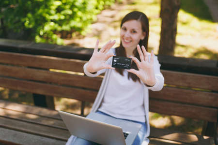 Young successful businesswoman holding credit card. Woman sitting on bench working on modern laptop...