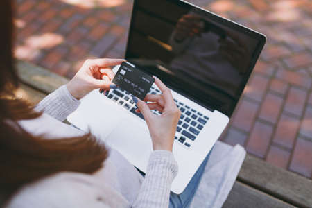 Close up female hands on keyboard. Woman sitting on bench holding credit card, working on modern laptop pc computer with blank empty screen outdoors. Mobile Office. Online shopping, business conceptの写真素材