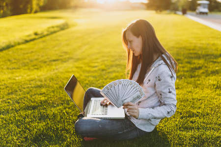 Female holding bundle of dollars cash money. Woman sitting on grass ground, working on laptop pc...