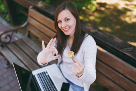 Young female in light casual clothes. Woman sitting on bench holding bitcoin coin of golden color working on modern laptop pc computer in street outdoors. Mobile Office virtual currency conceptの写真素材