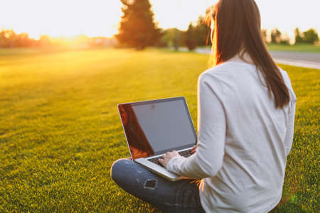 Young female back view. Woman working on laptop pc computer with blank black empty screen to copy space in city park on green grass sunshine lawn outdoors. Mobile Office. Freelance business conceptの写真素材