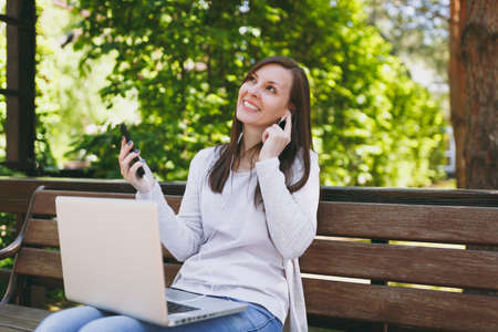 Young girl listen music in mobile phone with earphones. Woman sitting on bench working on modern laptop pc computer in city park in street outdoors on nature. Mobile Office Freelance business conceptの写真素材