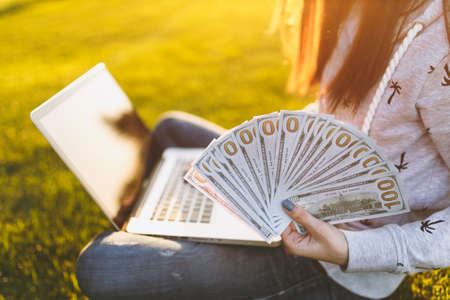 Female holding bundle of dollars cash money. Woman sitting on grass ground, working on laptop pc...
