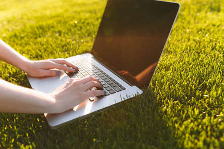 Close up hands on keyboard. Woman working on laptop pc computer with blank black empty screen to copy space in park on green grass sunshine lawn outdoors. Mobile Office. Freelance business conceptの写真素材