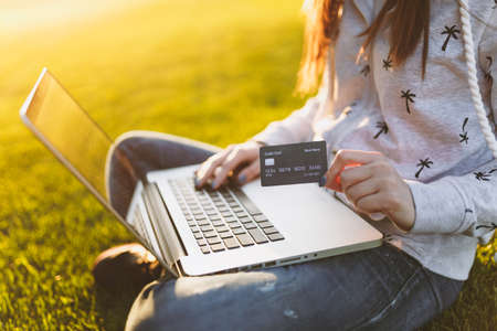 Young happy student female with credit card. Woman sitting on grass ground, working on laptop pc computer in city park on green grass sunshine lawn outdoors. Mobile Office. Freelance business conceptの写真素材