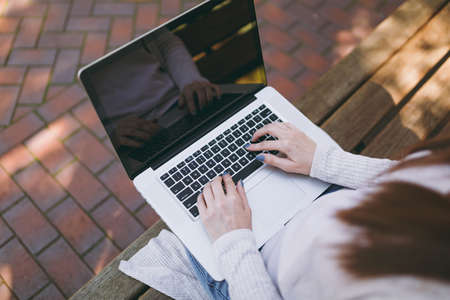 Close up cropped successful female hands on keyboard. Woman sitting on bench working on modern laptop pc computer with blank empty screen in street outdoors. Mobile Office Freelance business conceptの写真素材