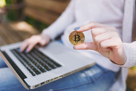 Close up female hands on keyboard. Woman sitting on bench holding bitcoin, coin of golden color,...