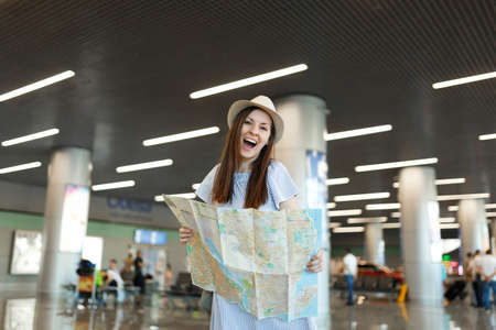 Young funny traveler tourist woman in hat holding paper map, searching route while waiting in lobby...
