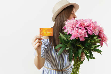 Young happy woman in blue dress, hat holding credit bank card, money, bouquet of beautiful pink peonies flowers isolated on white background. Business, delivery, online shopping concept. Copy spaceの写真素材