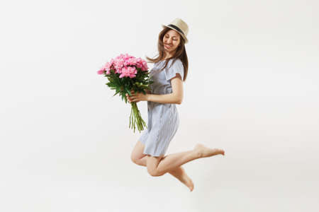 Full length of happy fun young woman in blue dress, hat holding bouquet of pink peonies flowers, jumping isolated on white background. St. Valentine's Day, International Women's Day holiday conceptの写真素材