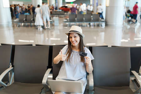 Young traveler tourist woman working on laptop, hold credit card, showing thumb up, waiting in lobby...