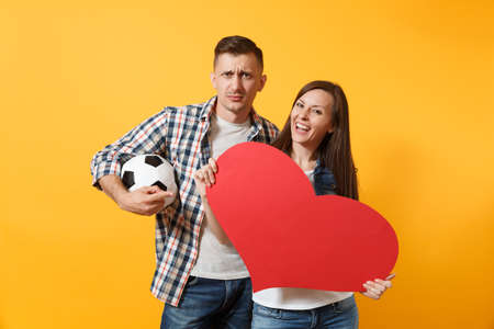 Young happy cheerful couple supporter, woman man, football fans cheer up support team, holding red heart love, soccer ball isolated on yellow background. Sport, family leisure, lifestyle conceptの写真素材