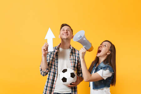 Happy fun expessive couple woman man football fans screaming, cheer up support team with soccer ball, white up arrow, megaphone isolated on yellow background. Sport family leisure lifestyle conceptの写真素材