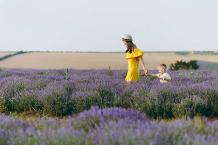 Young woman in yellow dress walk on purple lavender flower meadow field background, rest, have fun, play with little cute child baby boy. Mother, small kid son. Family day, parents, children conceptの写真素材