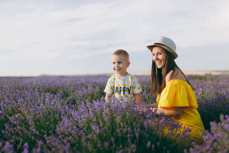 Young woman in yellow dress walk on purple lavender flower meadow field background, rest, have fun, play with little cute child baby boy. Mother, small kid son. Family day, parents, children conceptの写真素材