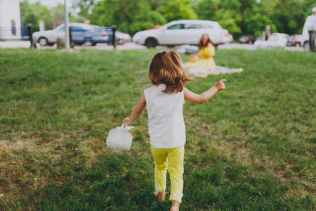 Back view of little cute child baby girl with basket running, play and have fun on green grass lawn...