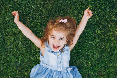 Laughing little pretty child baby girl in light dress lie on green grass lawn, spreading hands in park. Mother, little kid daughter. Mother's Day, love family, parenthood, childhood conceptの写真素材