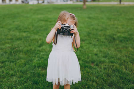 Portrait of little cute child baby girl in light dress holding retro vintage photo camera on green grass in park. Mother, little kid daughter. Mother's Day, love family, parenthood, childhood conceptの写真素材