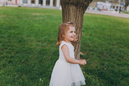 Laughing little cute child baby girl in light dress stand and lean on tree on green grass in city...