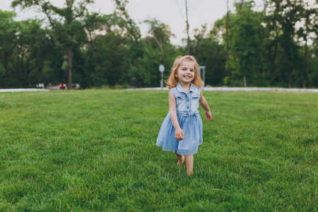 Smiling little cute child baby girl in denim dress walking and running, have fun on green grass lawn in park. Mother, little kid daughter. Mother's Day, love family, parenthood, childhood conceptの写真素材