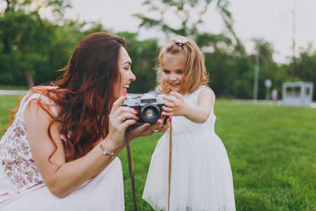Laughing woman in light dress and little pretty child baby girl holding retro vintage photo camera in green park. Mother, little kid daughter. Mother's Day, love family, parenthood, childhood conceptの写真素材