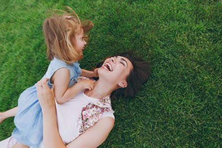 Overjoyed woman in light dress and little cute child baby girl lie on green grass in park rest play...